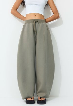 Femme portant un pantalon de jogging large vert olive, un débardeur court blanc et des sandales à plateforme noires, debout sur fond blanc.