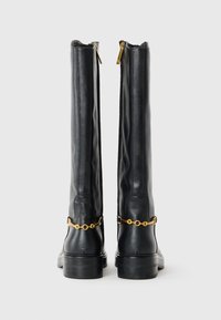 Bottes hautes en cuir noir avec un détail de chaîne dorée au-dessus du talon et des fermetures éclair arrière visibles sur un fond clair.