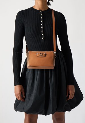 Sac bandoulière en cuir marron avec ferrures argentées, détails cousus et forme rectangulaire. Porté sur un haut noir côtelé et une jupe volumineuse.