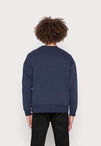 Person med lockigt hår som bär en enkel marinblå sweatshirt och svarta byxor, stående med ryggen mot en vit bakgrund.