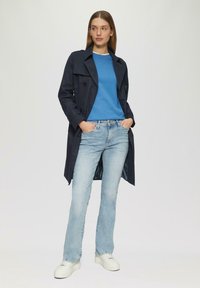 Marineblauer Trenchcoat mit doppelreihigen Knöpfen, getragen über einem blauen Pullover und hellblauen Jeans. Hat einen weißen gestreiften Innenfutter und weiße Sneakers.