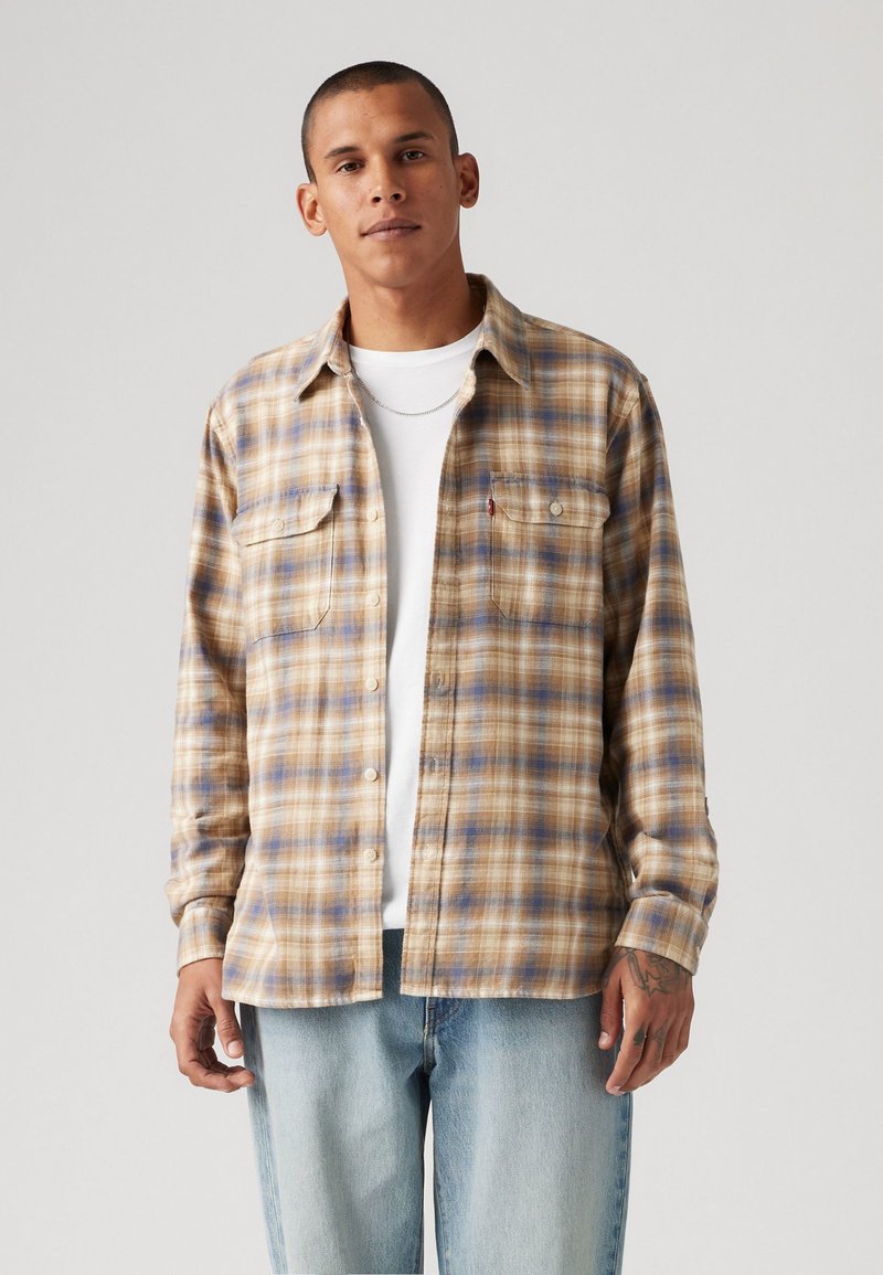 Camisa de flanela xadrez em tons de bege e azul, com dois bolsos no peito e fecho de botões. Usada por cima de uma t-shirt branca e jeans azul claro.