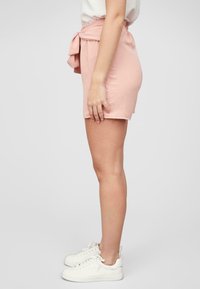 Shorts roses en tissu doux, avec un détail de taille nouée, longueur mi-cuisse et texture lisse. Portés avec des baskets blanches.