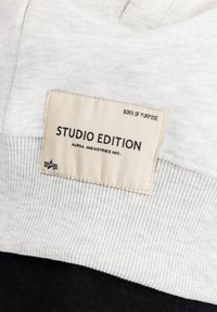 Osvetljen sivi ovratnik dukserice z rebrasto teksturo. Kremna tkaninska oznaka z napisom "STUDIO EDITION" in pod njim "ALPHA INDUSTRIES INC.".