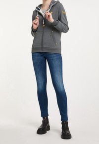 Sudadera gris con cremallera y capucha forrada, detalles en amarillo y bolsillos frontales, combinada con jeans ajustados azules y botas negras hasta los tobillos.