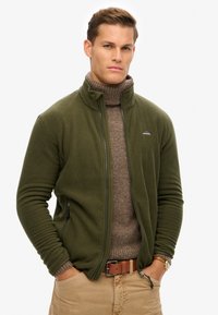 Zielona oliwkowa polarowa kurtka z zamkiem z przodu, wysokim kołnierzem, bocznymi kieszeniami i ściągaczami na mankietach. Nosi się ją na brązowym swetrze z golfem.