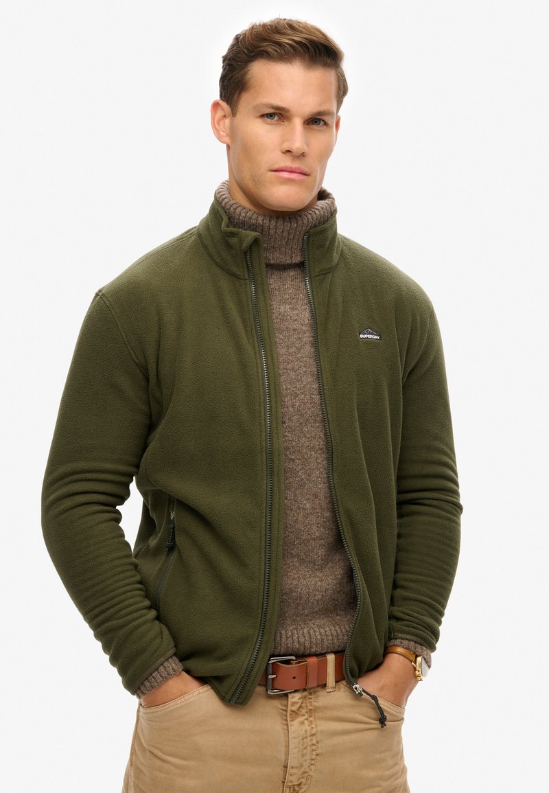 Veste en polaire vert olive avec fermeture éclair sur le devant, col haut, poches latérales et poignets côtelés. Portée par-dessus un pull à col roulé marron.