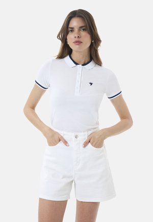 Polo bianco a maniche corte, colletto blu navy e rifiniture delle maniche blu navy, con un piccolo logo a forma di uccello. Abbinato a pantaloni corti in denim bianchi.