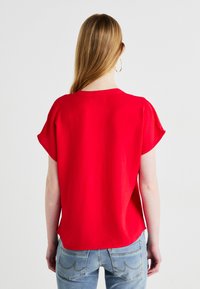 Top rosso a maniche corte con vestibilità rilassata, caratterizzato da una texture morbida e un design posteriore casual. Abbinato a jeans blu.