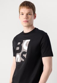 Ung mann med kort lys brun hår iført en svart T-skjorte med store hvite og mønstrede abstrakte bokstaver på forsiden.
