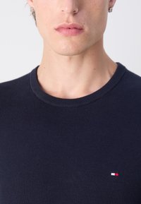 Marineblauer Strickpullover mit Rundhalsausschnitt und gerippten Bündchen, versehen mit einem kleinen rot-weiß-blauen Logo auf der Brust. Glatte Textur.