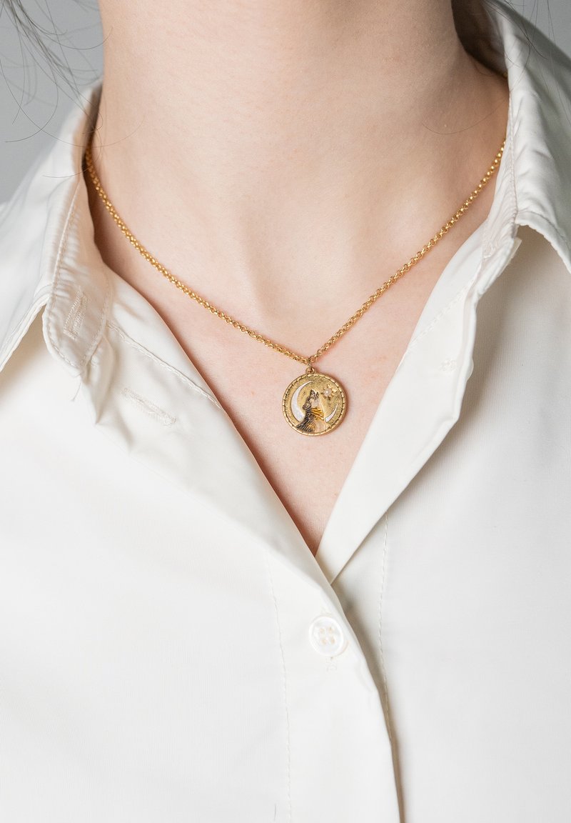 Guldsmycke med ett runt hänge som har en katt-silhuett, månskära och stjärna, buren av en person med en vit skjorta med knappar.