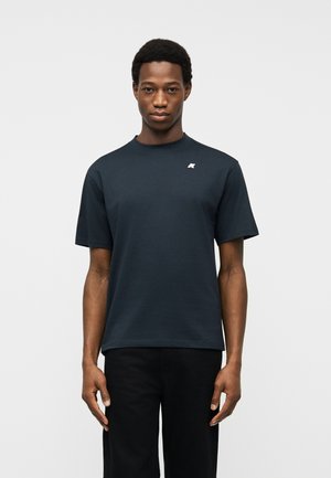 Hombre que lleva una camiseta lisa de manga corta azul marino oscuro con un pequeño logo blanco en el pecho izquierdo, de pie frente a un fondo gris claro.