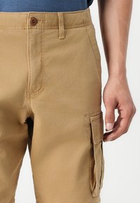 Khaki spodnie cargo mają zapięcie na guzik, kieszeń boczną oraz teksturowany materiał. Projekt obejmuje prosty krój z dokładnym wykończeniem szwów.