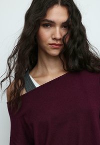 Bordeauxroter, schulterfreier Pullover mit weicher Textur und lockerem Schnitt. Darunter sind graue und weiße, geschichtete Oberteile sichtbar.