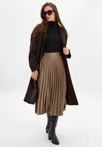 Brauner Wollmantel über einem schwarzen Rollkragenpullover, kombiniert mit einem plissierten beige Rock und schwarzen knielangen High Heels. Dunkle übergroße Sonnenbrille.