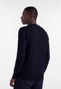 Mornarska pulover z teksturiranim mrežastim vzorcem, rebrastimi manšeti in robom ter klasičnim okroglim izrezom, oblečen z temnim trdim jeansom.