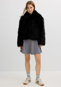 Mujer vestida con una chaqueta de piel sintética negra, falda gris, calcetines grises y zapatillas blancas, de pie contra un fondo claro y liso.