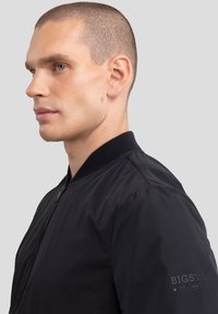 Juodas bombonešis su užtrauktuku priekyje, raštuota apykakle ir elastingomis rankovėmis. Ant rankovės yra logotipas, švelni tekstūra ir minimalus dizainas.