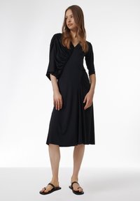 Vestido negro midi con detalles drapeados en un hombro, escote en V, silueta fluida y mangas cortas. Llevado con chanclas negras.