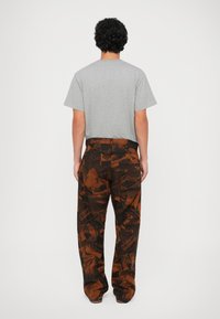 Graues Kurzarm-T-Shirt kombiniert mit locker sitzenden braunen Hosen, die ein schwarzes und oranges abstraktes Muster und sichtbare Gesäßtaschen aufweisen.