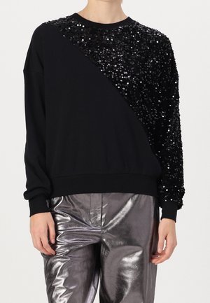 Kobieta ubrana w czarną bluzę z ukośnymi czarnymi cekinami na ramieniu oraz błyszczące metaliczne srebrne spodnie, stojąca z rozluźnionymi rękami.