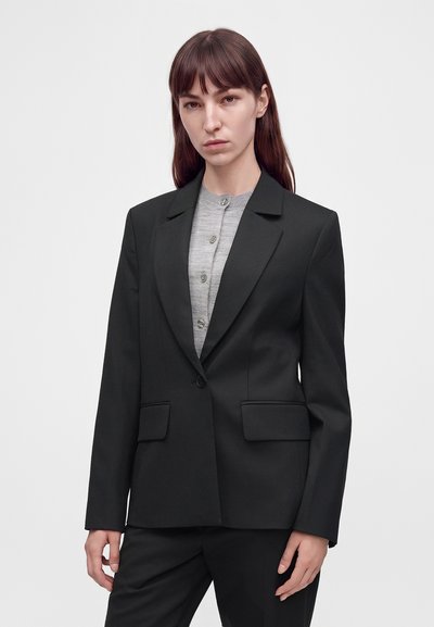 Femme aux cheveux bruns et raides portant un blazer noir cintré sur une chemise gris clair boutonnée, sur un fond clair uni.