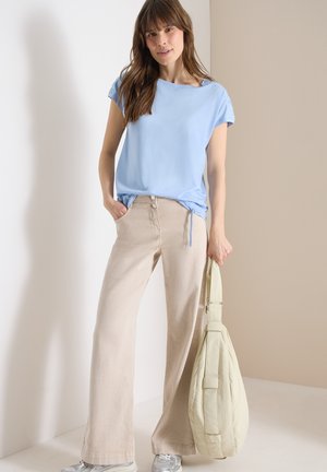 Femme portant un haut bleu clair et un pantalon ample beige, tenant un sac matelassé crème dans une pièce aux tons neutres.