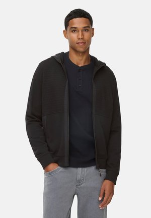 Hoodie noir à zip avec des panneaux texturés, associé à une chemise henley marine. Le mannequin porte un jean gris. Tissu lisse, coupe décontractée.