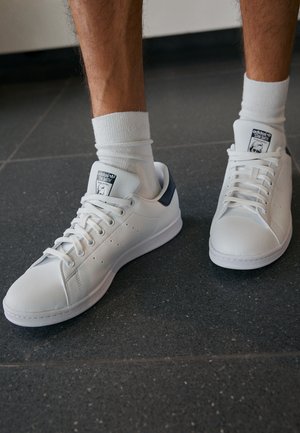 Hvide lædersneakers med en glat finish, der har en blå hælaccent og perforerede detaljer. Snørebånd og mærketags synlige.