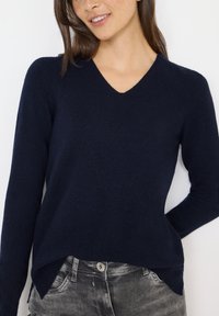 Marineblauer V-Ausschnitt-Pullover aus weich strukturiertem Strickstoff. Hat lange Ärmel und einen leicht asymmetrischen Saum. Kombiniert mit grauen Jeans.