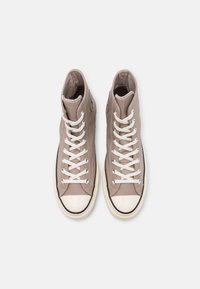 Wysokie trampki w jasnym taupe z płótna, z białymi sznurówkami, gumowym czubkiem, oraz charakterystyczną naszywką logo, cechujące się przeszyciami i teksturowanym wykończeniem.