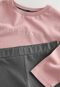 Ružičasti pamučni majica sadrži srebrni natpis "ALLSAINTS", u kombinaciji sa sivoj pamučne šortsevima, prizor koji prikazuje glatku teksturu i opušteni kroj.