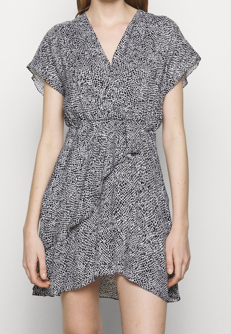 Femme portant une robe portefeuille courte à pois noirs et blancs, avec des manches courtes et un ourlet à volants.
