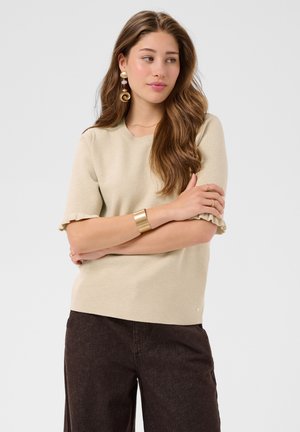 Jeune femme aux longs cheveux bruns portant un haut beige à manches courtes, un pantalon foncé, un large bracelet en or et des boucles d'oreilles décoratives, debout, les bras croisés.