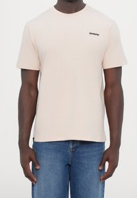 Camiseta de algodón color rosa claro con mangas cortas, cuello redondo y pequeño logo negro "BARROW" en el lado izquierdo del pecho. Combinada con vaqueros azul denim.