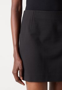 Gros plan d'une personne portant une jupe noire unie et un haut noir, montrant le bras gauche et la main détendus sur le côté.