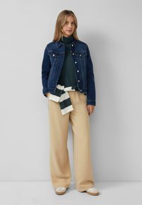 Dunkelblaue Jeansjacke; langärmliger dunkelgrüner Rollkragenpullover; beigen weit geschnittene Hose; gestreifte Krawatte in der Taille; hellfarbene Schuhe.