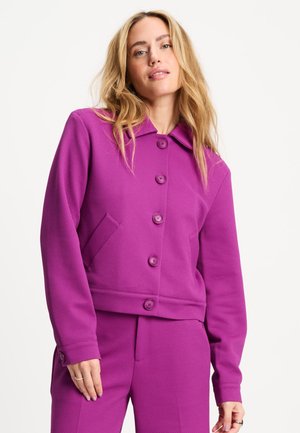 Vrouw met lang blond haar die een gestructureerde paarse button-up jas en bijpassende broek draagt tegen een effen achtergrond.