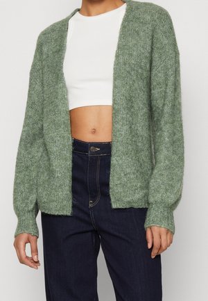 Osoba ubrana w zielony, otwarty sweter z dzianiny, na białym krótkim topie i ciemnoniebieskich, wysokich jeansach, stojąca z rozluźnionymi rękami.