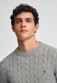 Grauer Kabelstrickpullover mit Rundhalsausschnitt und gerippten Bündchen, mit einem kleinen Logo-Akzent auf der Brust und strukturiertem Stoffmuster.