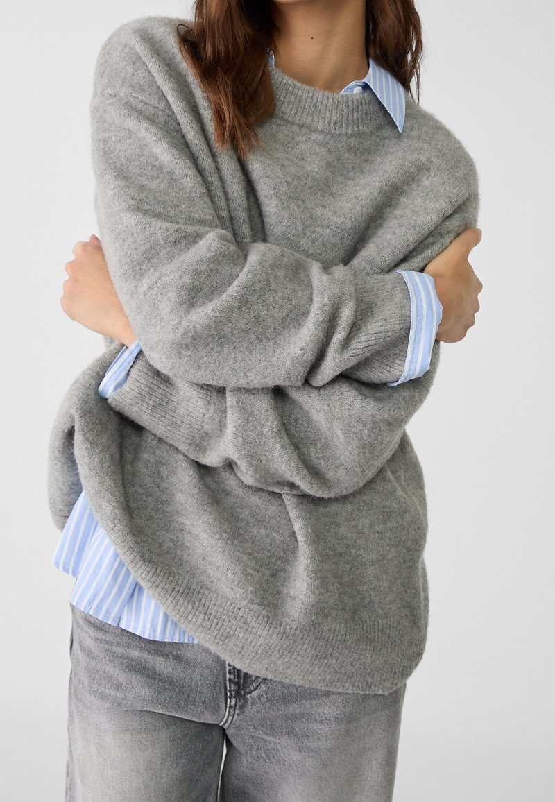 Personne portant un pull en laine gris par-dessus une chemise rayée bleu et blanc, les bras croisés, associée à un jean gris sur un fond uni.