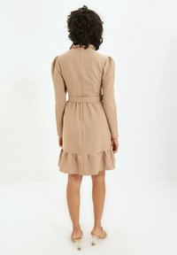 Beiges Kleid mit langen Ärmeln, tailliertem Oberteil und Rüschensaum. Hergestellt aus glattem Stoff mit tailliertem Bund und Reißverschluss auf der Rückseite.