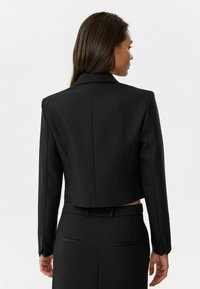 Zwart cropped blazer met een getailleerd ontwerp, voorzien van scherpe revers, gestructureerde schouders en knoopaccenten op de mouwen.