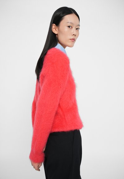 Maglione morbido e rosso brillante con una vestibilità rilassata, maniche lunghe e polsini a costine, indossato sopra una camicia colletto azzurra e pantaloni neri.