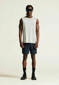 Atletische man die staat, in een mouwloos grijs shirt, marineblauwe shorts, zwarte sokken, zwarte schoenen, blauwe visors zonnebril en een armband om zijn rechterpols.