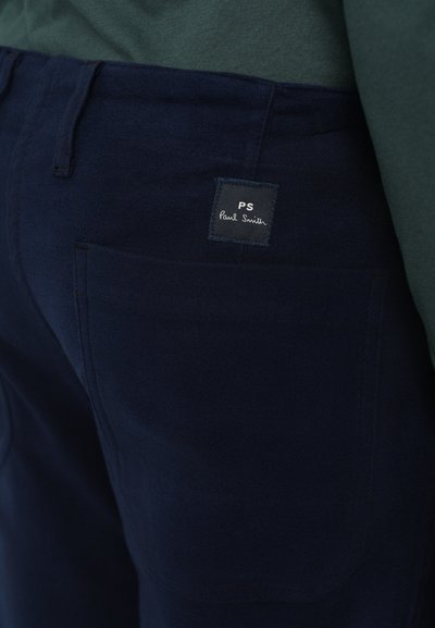 Mörkblå byxor med bakficka och svart fyrkantig Paul Smith logopatch, bärs med en mörkgrön topp.
