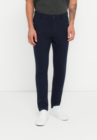 Pantaloni slim-fit blu navy con chiusura a bottone, tasche e una texture liscia, abbinati a sneaker bianche.