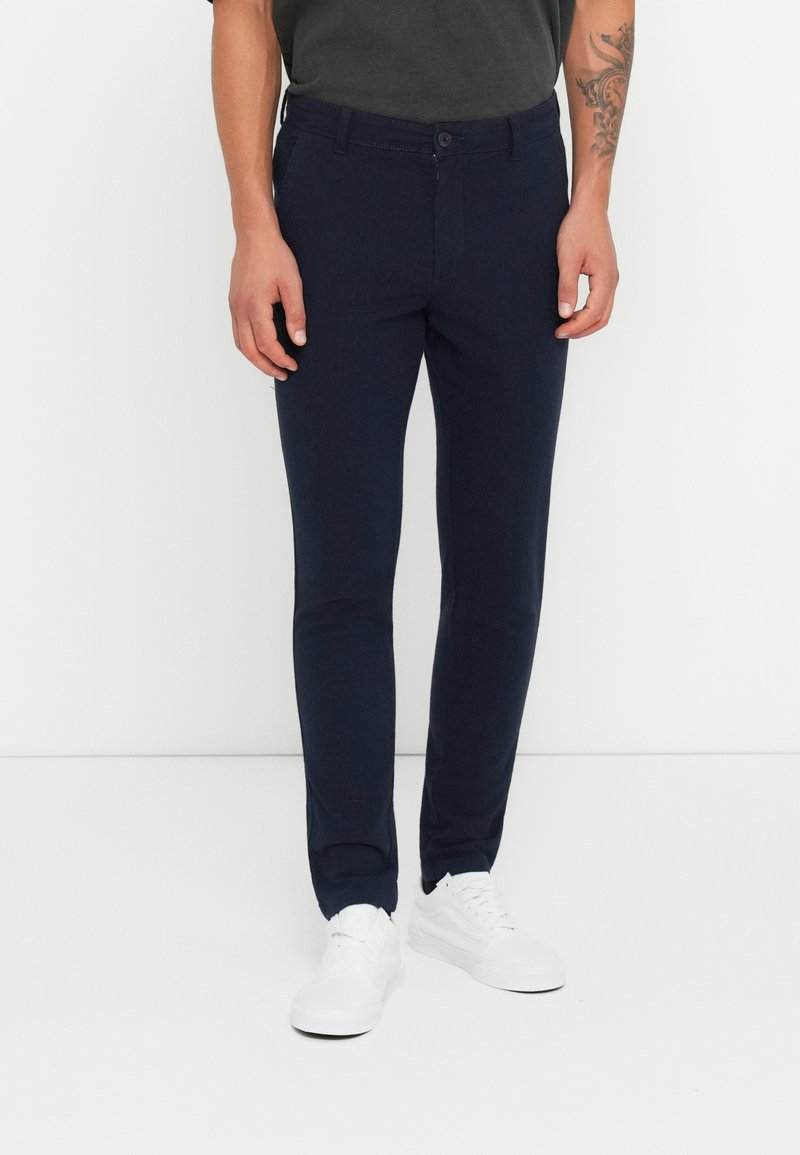 Pantaloni slim-fit blu navy con chiusura a bottone, tasche e una texture liscia, abbinati a sneaker bianche.