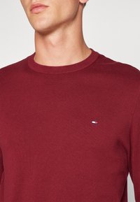 Pull en maille côtelée rouge avec un col rond et un petit logo brodé en bleu, blanc et rouge sur le côté gauche de la poitrine. Texture douce.